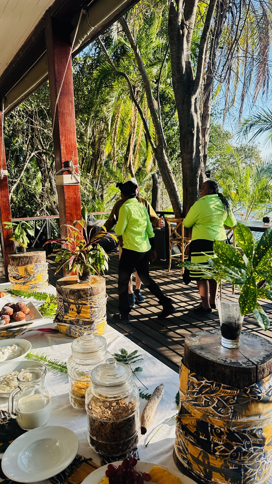 Breakfast laid out on the lodge deck under palms beside the river, with staff in green
